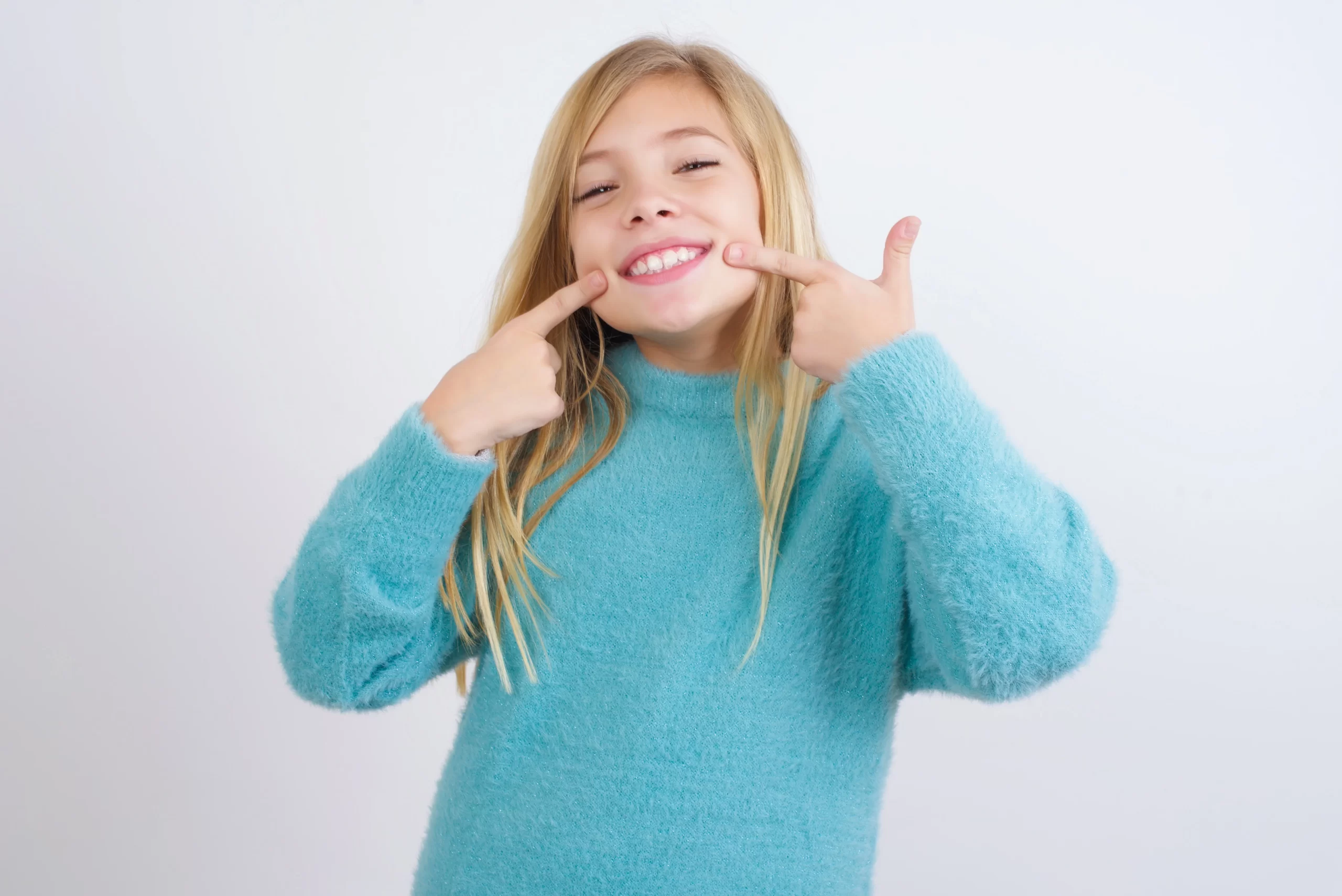 Smiling child in blue sweater points at teeth.