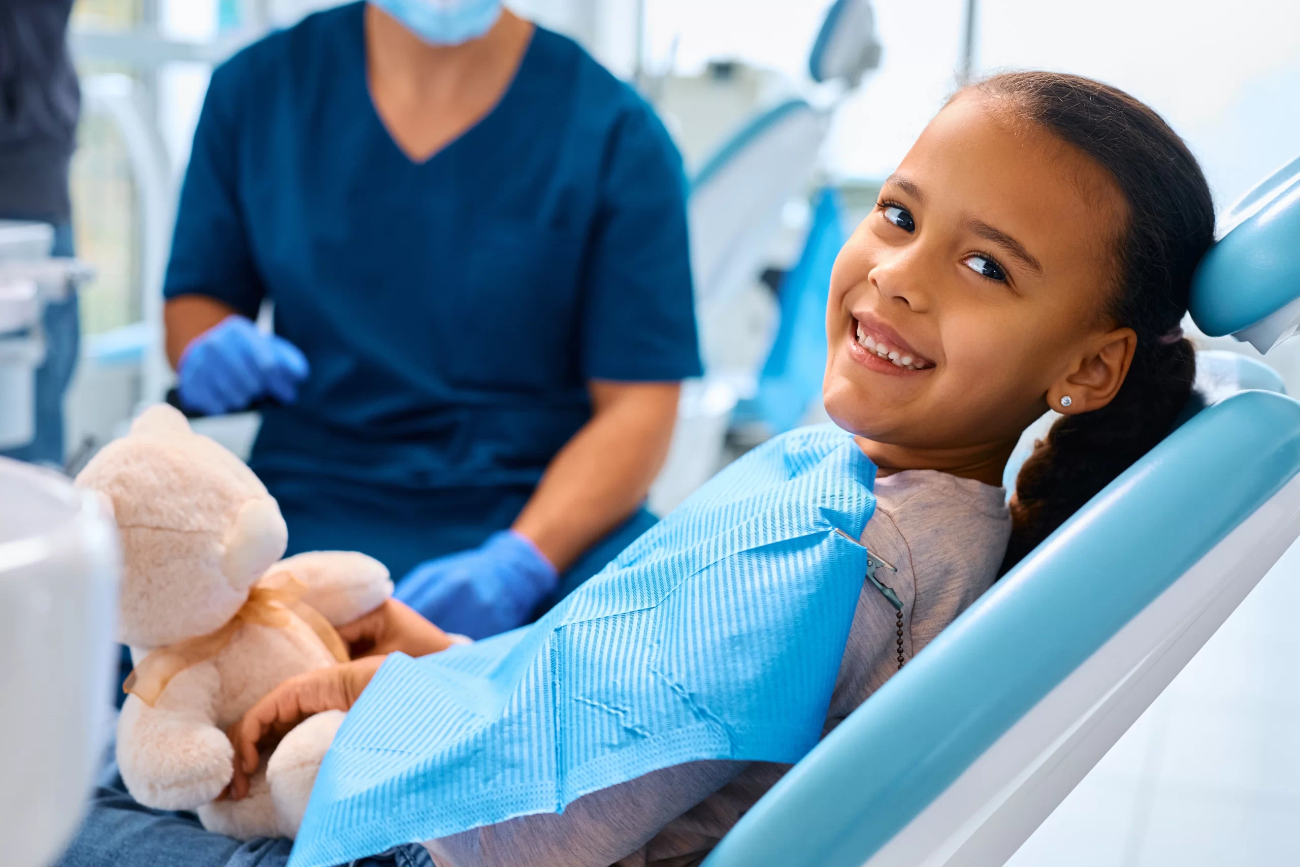 Child smiling in dental chair with teddy bear.